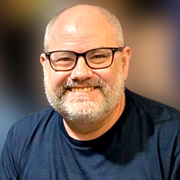 A professional, warm headshot of Matthew Saunders, the AI Ambassador for amazee.io and amazee.ai, and a key leader in the Drupal AI Initiative. He is smiling, wearing black-rimmed glasses and a dark blue t-shirt, captured against a softly blurred, neutral-colored background.